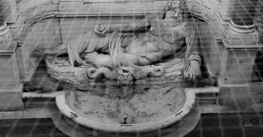 Black and white photo of an ancient marble statue resembling a reclining Greek or Roman god in an ornate fountain.